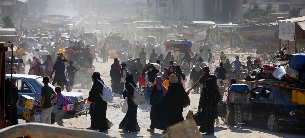 Civiles palestinos caminan por una calle polvorienta de Gaza, cargando sus pertenencias entre edificios dañados y escombros.