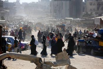 Civiles palestinos caminan por una calle polvorienta de Gaza, cargando sus pertenencias entre edificios dañados y escombros.