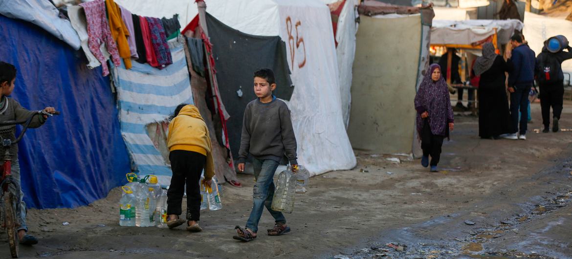 A young boy carries a water bottle through an area where people are living in tents.