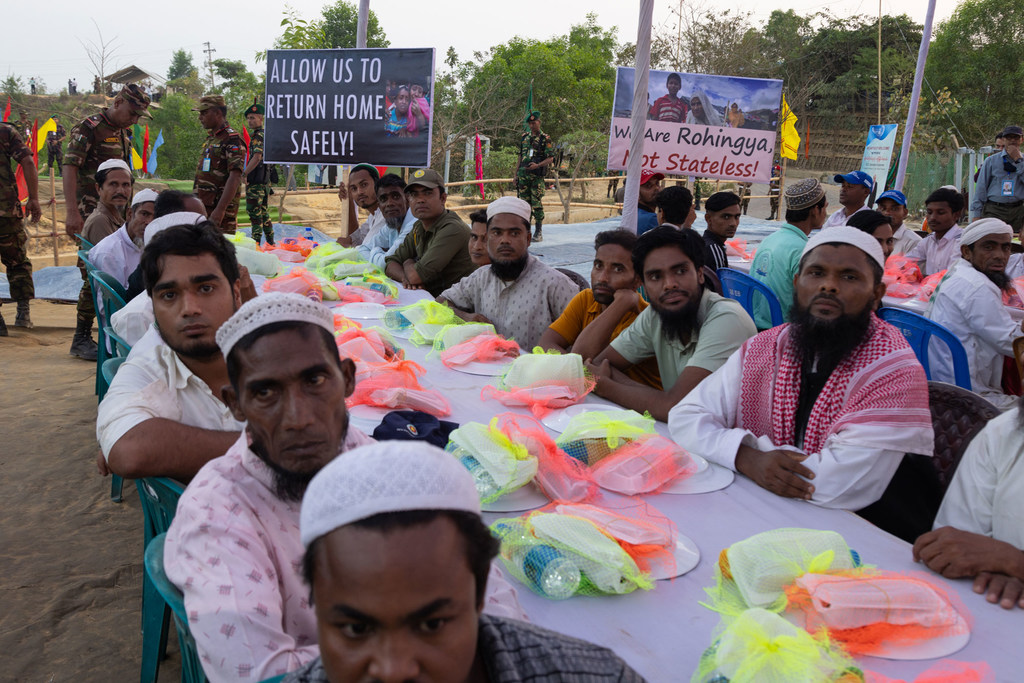 Kundi la wakimbizi wa Rohingya nchini Bangladesh wameketi kwenye meza ndefu wakiwa na vifaa, wakiwa na mabango yenye maandishi 'TURUHUSU KUREJEA NYUMBANI SALAMA!' na 'Sisi ni Warohingya, Sio Wasio na Uraia!'