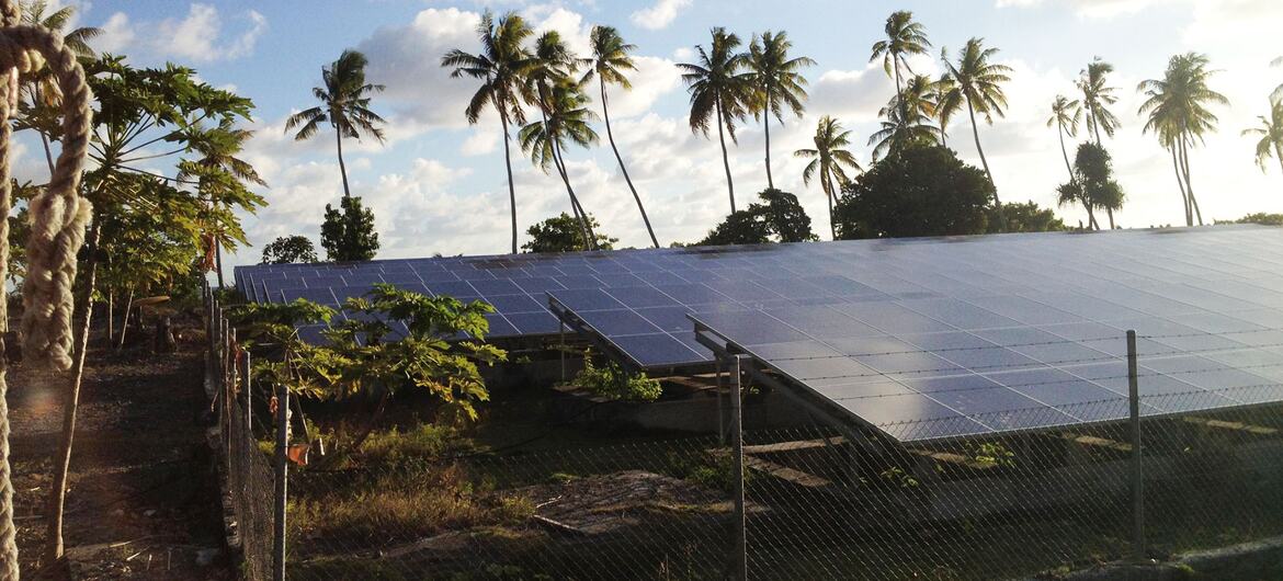 Paneles solares instalados en una parcela cercada en Tokelau, un grupo de tres atolones en el Pacífico Sur, con palmeras en el fondo bajo un cielo azul con nubes blancas.