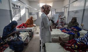 MSF nurse Maha stands in a maternity ward in Tawila, North Darfur, Sudan, reviewing patient records while surrounded by displaced mothers and newborns on hospital beds. The photo captures the healthcare challenges in the Darfur displacement crisis.