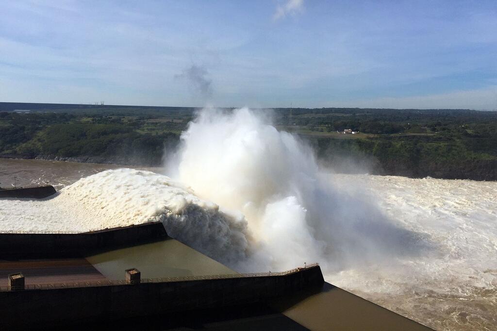 إطلاق هائل للمياه من سد إيتايبو على نهر بارانا، مما يُحدث سحابة ضخمة من المياه البيضاء والرذاذ ضد سماء زرقاء. يُدار السد بشكل مشترك من قبل البرازيل وباراغواي.