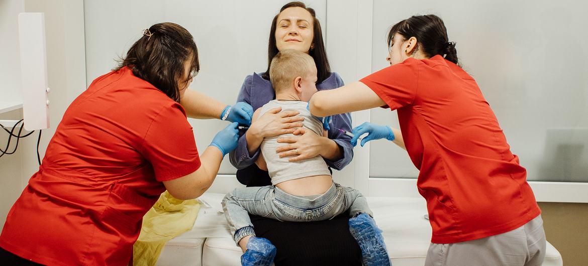 Two nurses in red scrubs administer a vaccination to a young boy held by his mother during a clinic visit.