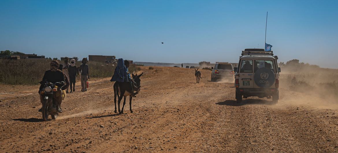 Um veículo de comboio do Ocha e uma motocicleta trafegam por uma estrada poeirenta em El Geneina, Darfur Ocidental, Sudão.