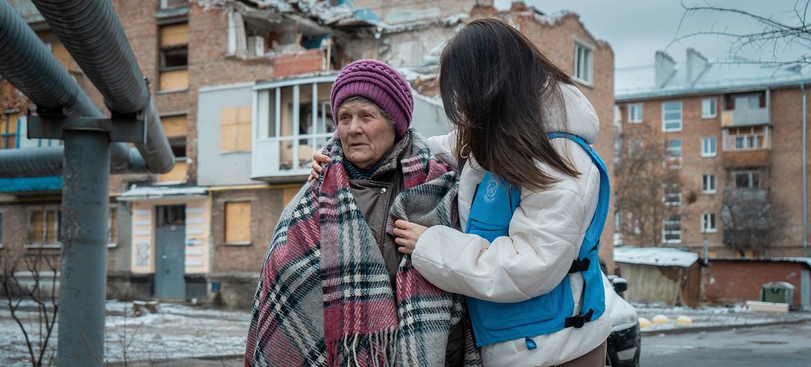 À Kharkiv, Olena, une Ukrainienne âgée, enveloppée dans une couverture à carreaux, se tient devant un immeuble endommagé. Un travailleur humanitaire en gilet bleu de OCHA la réconforte.