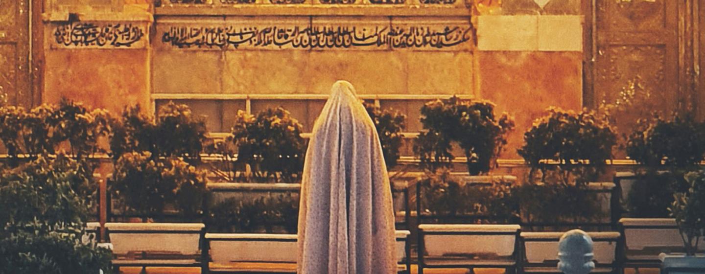 A woman wearing a chador stands on a stone courtyard with benches and Islamic calligraphy in the background.