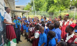 Anthurium Lewis, an archer from Tobago, speaks to a group of school children in uniform about the Sustainable Development Goals (SDGs), holding a small potted plant.