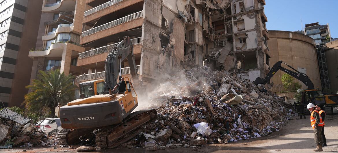 Heavy machinery clears rubble from a building destroyed by airstrikes in Beirut, Lebanon. Workers in safety gear are present at the site of the recent destruction.