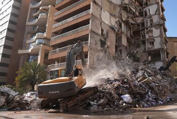 Heavy machinery clears rubble from a building destroyed by airstrikes in Beirut, Lebanon. Workers in safety gear are present at the site of the recent destruction.