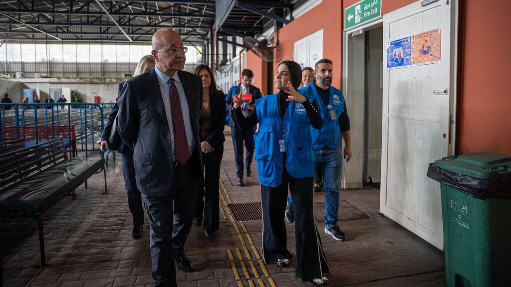 Le Haut-Commissaire des Nations Unies pour les réfugiés, Barham Salih, un homme chauve en costume et lunettes, est guidé à travers le Centre de traitement des dossiers du HCR à Jnah, Beyrouth, au Liban, par une agente du HCR portant un gilet bleu. D'autres agents et responsables du HCR les suivent dans un couloir où l'on aperçoit des bancs et un panneau de sortie de secours.