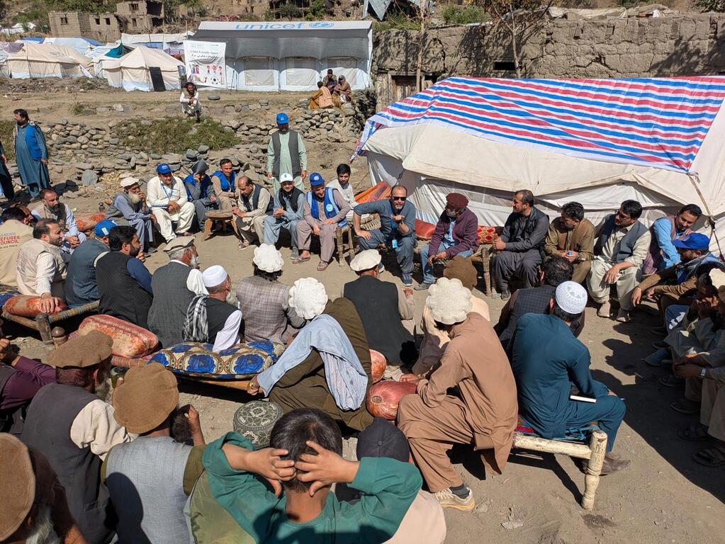 Representantes de ONU-Hábitat y UNICEF se reúnen con hombres afganos en un campamento de refugiados, con tiendas de campaña de la ONU visibles en el fondo.