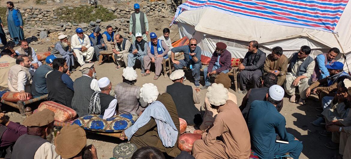 Representantes da ONU-Habitat e da UNICEF reúnem-se com homens afegãos em um campo de refugiados, com tendas da ONU visíveis ao fundo.