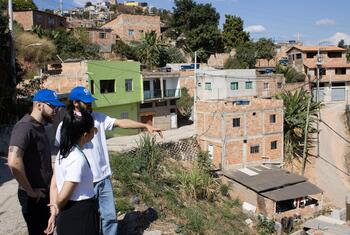 Three people in blue UN-Habitat caps stand on a hillside overlooking informal housing in Izidora, Belo Horizonte, Brazil, during a field visit.