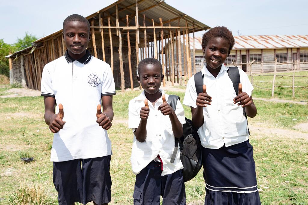Three school children in uniforms give thumbs up in front of a school building in Tchomia, DR Congo.