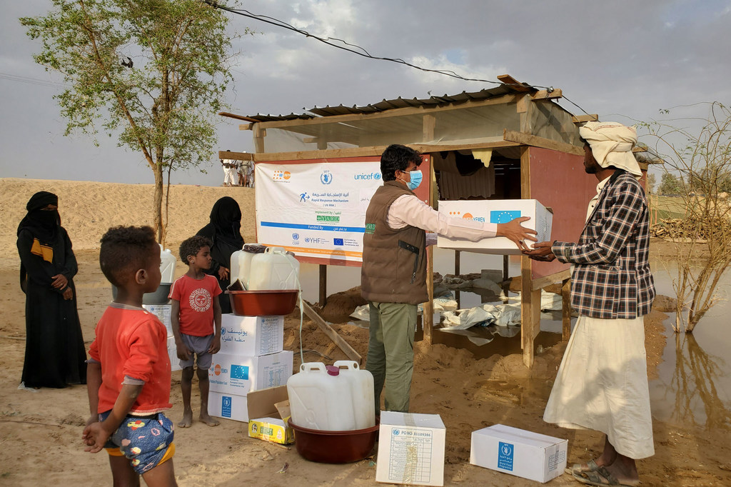 Un travailleur humanitaire de UNFPA et de UNICEF remet un carton de provisions à un homme dans un point de distribution du gouvernorat d'Al Jawf, au Yémen. Des enfants et des femmes observent la distribution des secours.