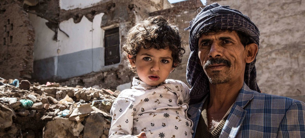 أب يحمل ابنته ويقف أمام منزله المدمّر في صنعاء.