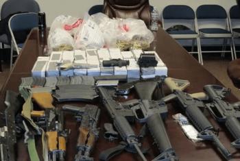 Una colección de armas y municiones ilegales incautadas colocadas en una mesa, incluyendo rifles de asalto, pistolas y cajas de municiones, en el contexto de la violencia de pandillas en Haití.