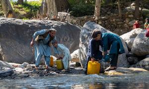 Afghan man and boys in traditional clothing collect water from a rocky mountain stream using yellow jerrycans after the earthquake displaced their family to temporary shelters in Kunar province.
