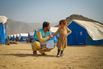 यूनिसेफ सीईएफसी सुविधा प्रदाता, नाजीरुल्लाह आदिल, अफगानिस्तान में एक विस्थापित शिविर में एक युवा लड़की के साथ बातचीत करने के लिए घुटने टेकते हुए, भूकंप की तैयारी के बारे में एक सुरक्षा ब्रोशर रखते हुए। नीले तम्बू और पहाड़ पृष्ठभूमि बनाते हैं।