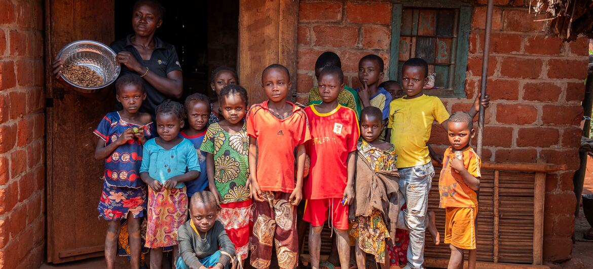 A group of children and a woman stand in front of a brick house in Bangassou, Central African Republic, during a press visit to assess the food crisis. The woman holds a bowl of grains, symbolizing emergency food assistance provided by the World Bank-funded PRUCAC project.