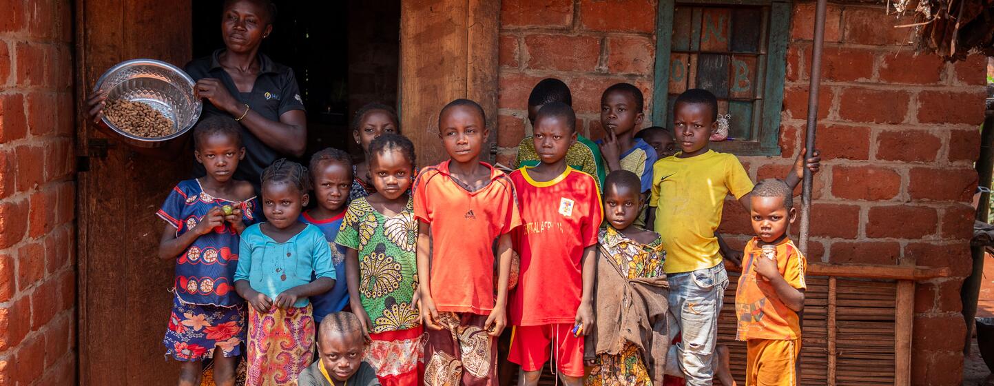 A group of children and a woman stand in front of a brick house in Bangassou, Central African Republic, during a press visit to assess the food crisis. The woman holds a bowl of grains, symbolizing emergency food assistance provided by the World Bank-funded PRUCAC project.