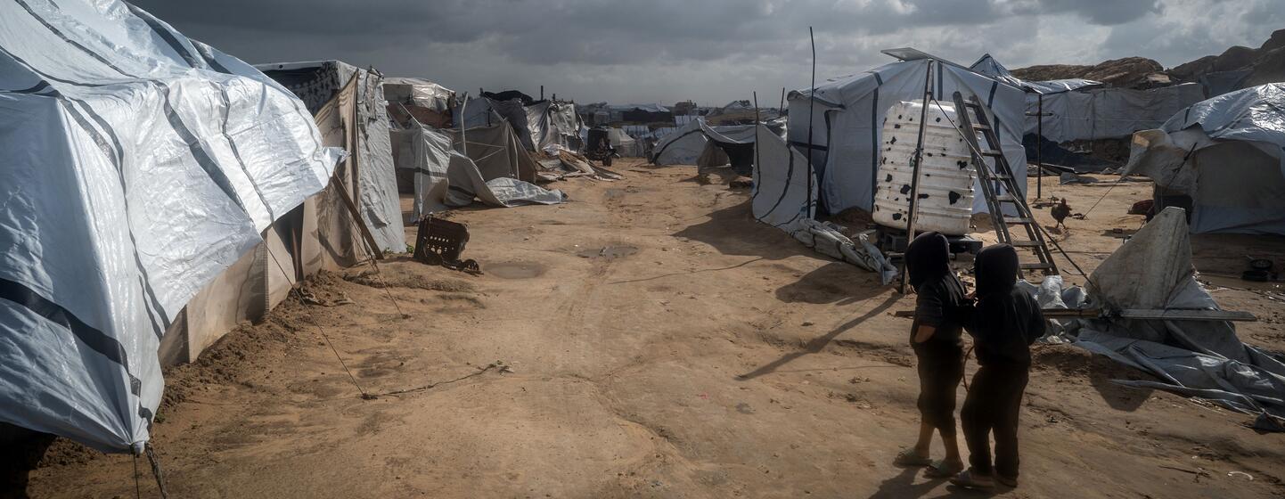 Deux enfants se tiennent dans un sentier poussiéreux entre des abris temporaires dans un camp de déplacés à Gaza, sous un ciel orageux.Les tentes sont visiblement endommagées par le vent et la pluie, soulignant les dures conditions de vie auxquelles sont confrontées les familles déplacées.