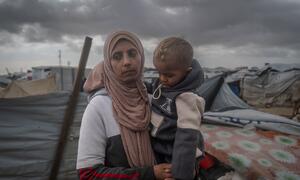 A Palestinian widow named Yasmin holds her young child in a makeshift tent shelter in Gaza. Her family was displaced by a storm that destroyed their tent and basic facilities, forcing her to send her children to relatives for safety. The image highlights the harsh winter conditions and lack of aid in coastal displacement sites.