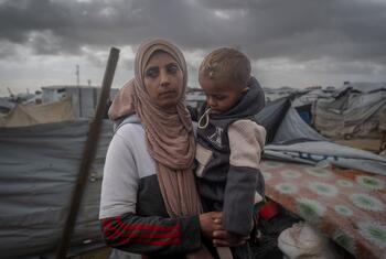 A Palestinian widow named Yasmin holds her young child in a makeshift tent shelter in Gaza. Her family was displaced by a storm that destroyed their tent and basic facilities, forcing her to send her children to relatives for safety. The image highlights the harsh winter conditions and lack of aid in coastal displacement sites.