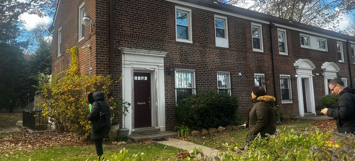 A photo of a red brick two-story townhouse on a residential street. Three people are visible walking on the sidewalk in front of the house. The scene is set in autumn with trees showing yellow and brown leaves.