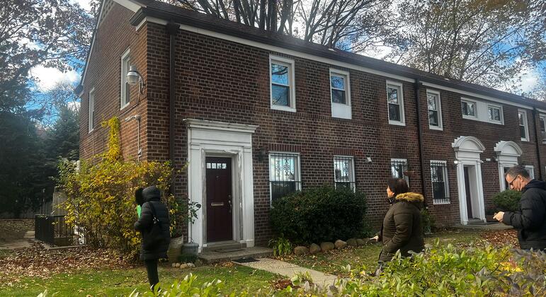 Фото красного кирпичного двухэтажного таунхауса на жилой улице. Три человека видны на тротуаре перед домом. Сцена разворачивается осенью, деревья показывают желтые и коричневые листья.