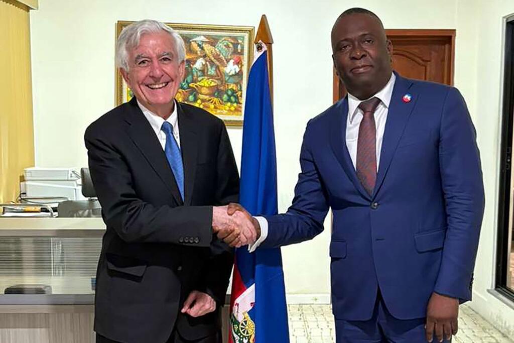 Une poignée de main formelle entre le diplomate français Jean-Yves Le Drian et le fonctionnaire haïtien Gabriel Pelissier dans un bureau, avec le drapeau haïtien en arrière-plan.