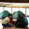 Четыре маленьких мальчика сидят в круге на полу большой комнаты, которая служит временным приютом. За ними - зеленые палатки для кемпинга, и на заднем плане видны другие люди. Это государственная школа в Бейруте, Ливан, преобразованная в приют для перемещенных из-за конфликта людей, где Всемирная продовольственная программа (ВПП) предоставляет горячие блюда.