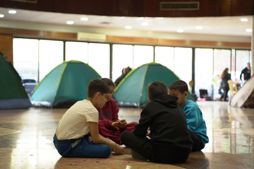 Quatre jeunes garçons sont assis dans un cercle sur le sol d'une grande pièce qui sert d'abri temporaire. Derrière eux sont des tentes de camping vertes, et en arrière-plan, d'autres personnes peuvent être vues. Ceci est une école publique de Beyrouth, au Liban, convertie en un refuge pour les personnes déplacées en raison du conflit, où le Programme alimentaire mondial (PAM) fournit des repas chauds.