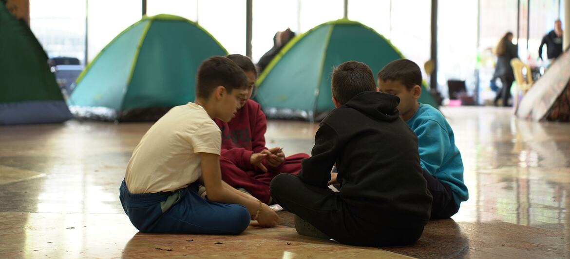 Four young boys sit in a circle on the floor of a large room that serves as a temporary shelter. Behind them are green camping tents, and in the background, other people can be seen. This is a public school in Beirut, Lebanon, converted into a shelter for displaced people due to conflict, where the World Food Programme (WFP) is providing hot meals.