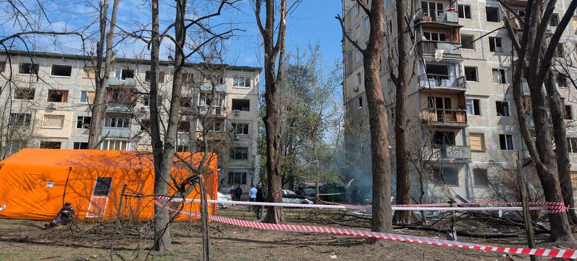 An orange emergency tent stands in front of a damaged apartment building in Ukraine after a missile and drone attack, with caution tape cordoning off the area and debris scattered on the ground.