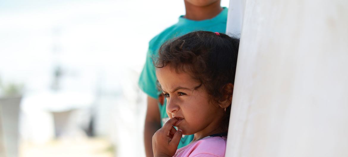 Une jeune fille aux cheveux bouclés, vêtue d'un t-shirt rose, se tient devant un mur blanc, le doigt dans la bouche. À l'arrière-plan, on aperçoit partiellement un garçon portant un t-shirt vert. La scène semble se dérouler en extérieur, sous un ciel lumineux.