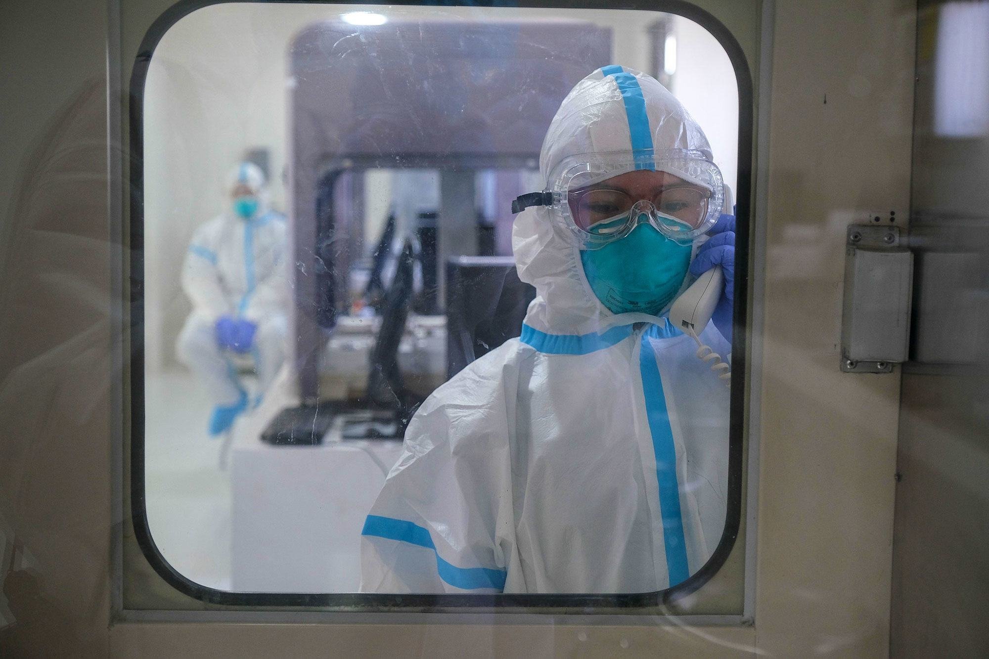 Un técnico médico, vestido con un traje protector blanco y azul completo y una careta facial, mira a través de una ventana mientras sostiene un teléfono en un entorno de laboratorio estéril.