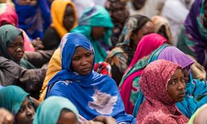 A group of Sudanese refugee women and children, wearing colorful headscarves, sit together in a refugee camp in Chad, highlighting the humanitarian crisis caused by the ongoing war in Sudan.