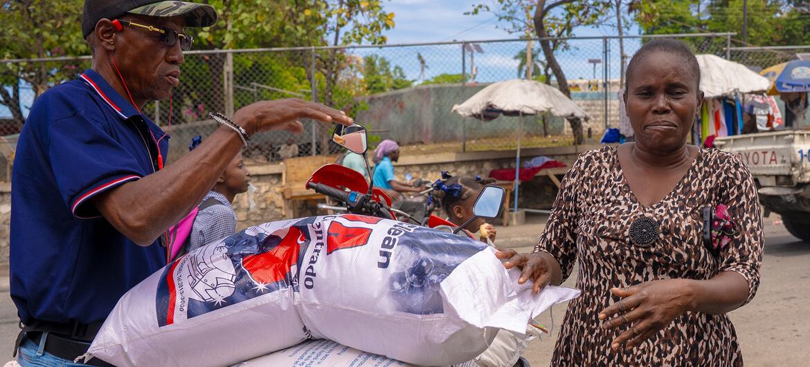 A man loads large sacks of rice onto a motorbike while a woman, identified as Dina, watches. This image depicts the World Food Programme (WFP) providing food assistance in Canapé Vert, Haiti.