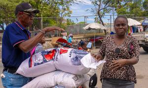 A man loads large sacks of rice onto a motorbike while a woman, identified as Dina, watches. This image depicts the World Food Programme (WFP) providing food assistance in Canapé Vert, Haiti.