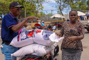 A man loads large sacks of rice onto a motorbike while a woman, identified as Dina, watches. This image depicts the World Food Programme (WFP) providing food assistance in Canapé Vert, Haiti.