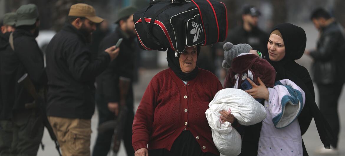 Two women carrying their belongings walk through a street in Syria, with armed men in the background.