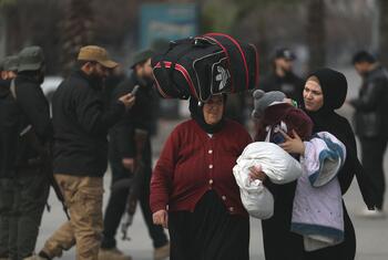Two women carrying their belongings walk through a street in Syria, with armed men in the background.