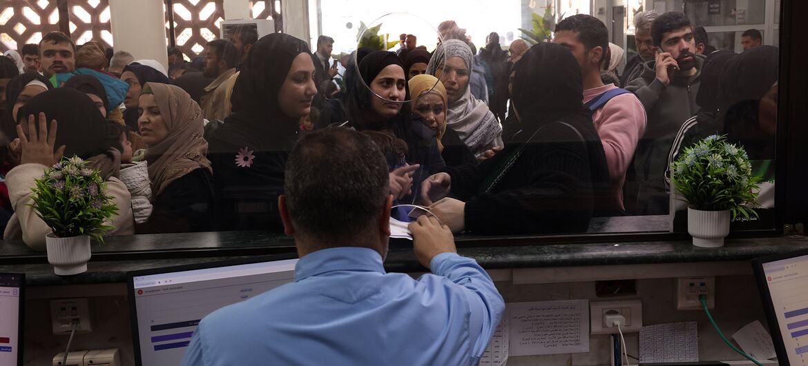 A crowded scene at an aid office where people, including women in hijabs and children, wait at a counter to receive assistance from staff.
