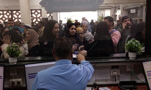 Una escena llena de gente en una oficina de ayuda donde personas, incluidas mujeres con hiyab y niños, esperan en un mostrador para recibir ayuda del personal.