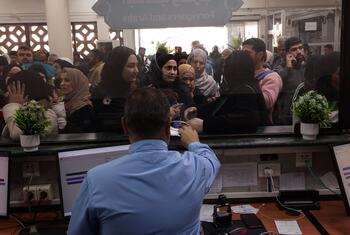 A crowded scene at an aid office where people, including women in hijabs and children, wait at a counter to receive assistance from staff.