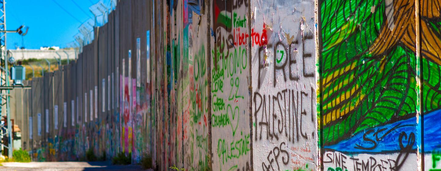 Une section de la barrière de séparation israélienne recouverte de graffitis colorés et de messages politiques, avec du fil barbelé visible au sommet sous un ciel bleu clair.