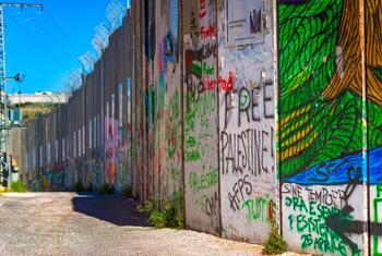 A section of the Israeli separation barrier covered in colorful graffiti and political messages, with barbed wire visible on top under a clear blue sky.