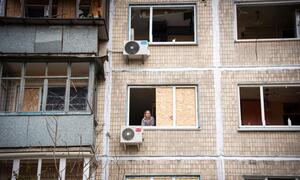 A woman looks out from a window of an apartment building with damaged windows and boarded-up panes in Kyiv, Ukraine.